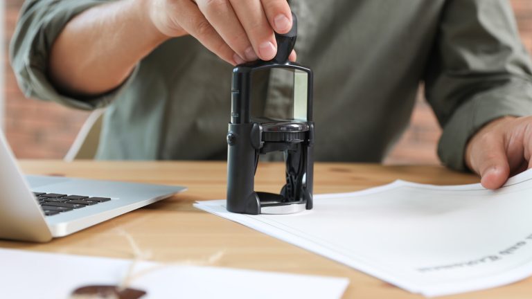 A hand pressing a self-inking stamp onto a sheet of paper on a desk beside a laptop